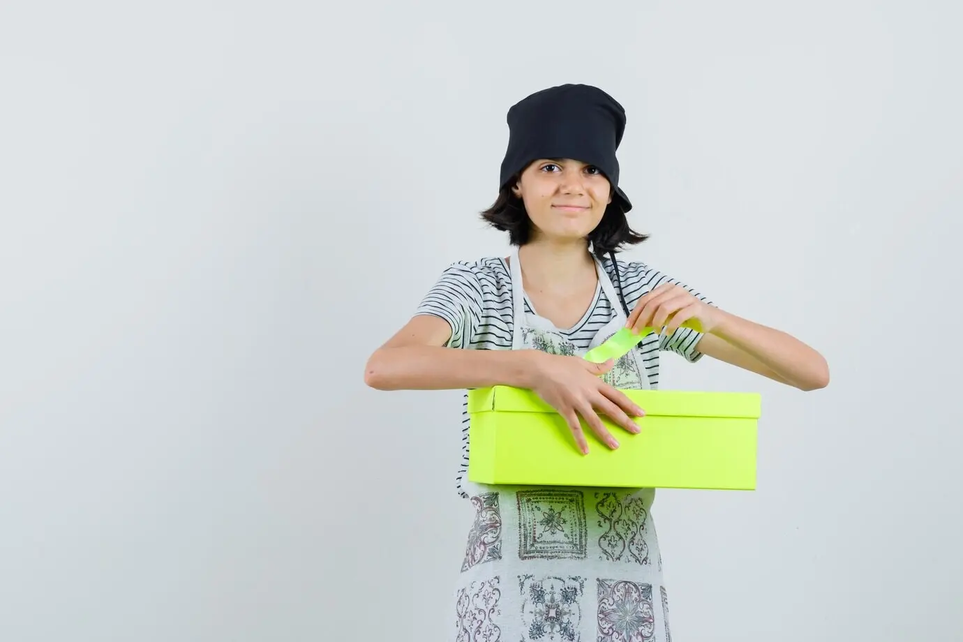 Маленькая девочка в кухонном платье открывает коробку с подарком и выглядит весёлой.